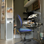 Libreria piccola bianca con libri e raccoglitori, scatola porta pipe appesa sotto e piano a ribalta grigio con computer MacMini e telefono del centralino interno dietro alla cassettiera grigia