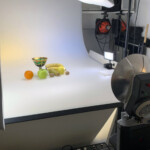 set fotografico con la composizione della frutta e del bicchiere, in lontananza due Ianiro arancioni al tungsteno